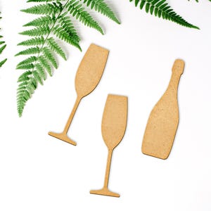 May include: Three wooden cutouts of a champagne bottle and two champagne glasses. The cutouts are on a white background.