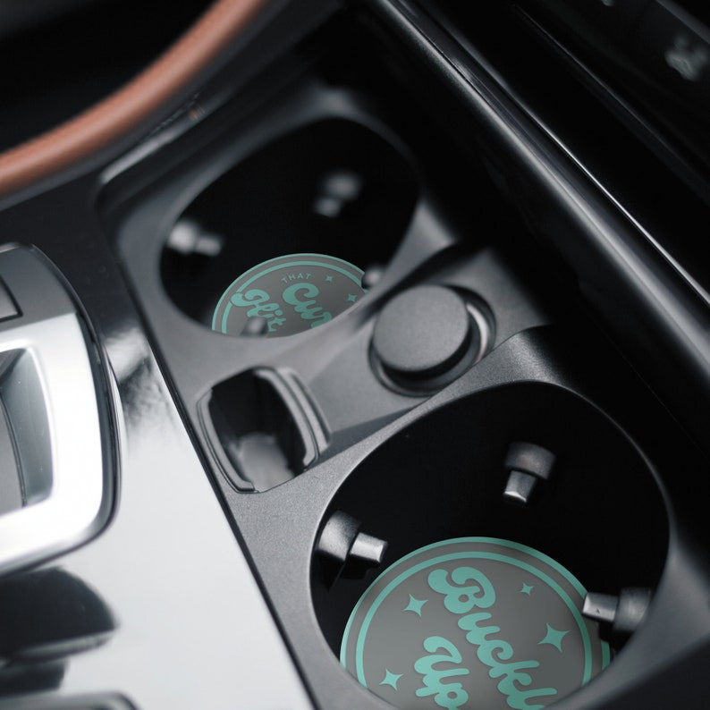 May include: Two round car coasters with a black background and teal text. The coaster on the left says "That Cup Life" and the coaster on the right says "Buckle Up".