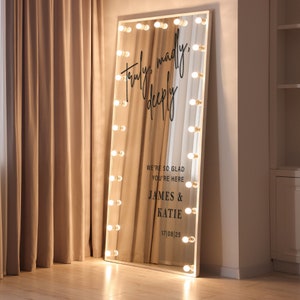 May include: A white vanity mirror with a white frame and a large mirror surface. The mirror has a row of light bulbs around the perimeter. The mirror has a black inscription that reads "Truly, madly, deeply" and "We're so glad you're here, James & Katie, 17/08/25".