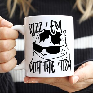 May include: White ceramic mug with a black and white illustration of a cat wearing sunglasses. The text "Rizz 'em with the 'tism" is printed on the mug.