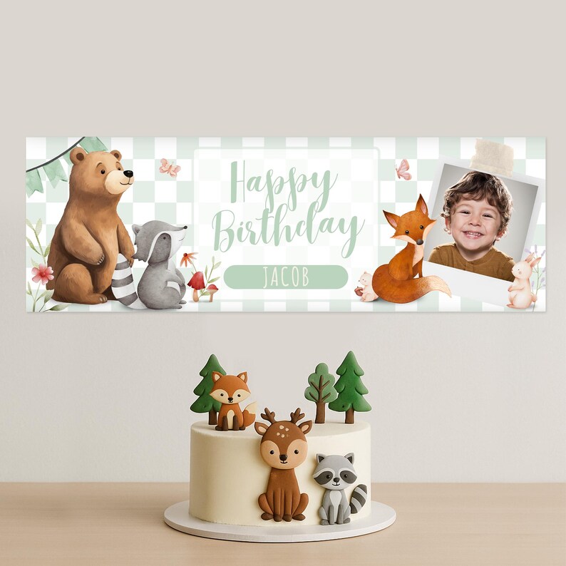 May include: A birthday banner with woodland animal illustrations and the text "Happy Birthday JACOB". A photo of a child is included. A cake with woodland animal cake toppers is in the foreground.