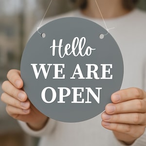 K&ouml;nnte beinhalten: Ein graues, rundes Schild mit den Worten "Hello WE ARE OPEN" in wei&szlig;er Schrift. Das Schild wird von einer Person gehalten. Das Schild hat zwei kleine L&ouml;cher oben zum Aufh&auml;ngen.