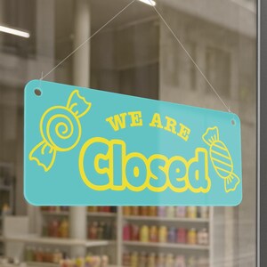 Puede incluir: Un letrero rectangular turquesa con esquinas redondeadas, colgado por un fino alambre. El letrero presenta las palabras "WE ARE Closed" en amarillo, junto con ilustraciones de caramelos amarillos a cada lado. El letrero se muestra en un escaparate.