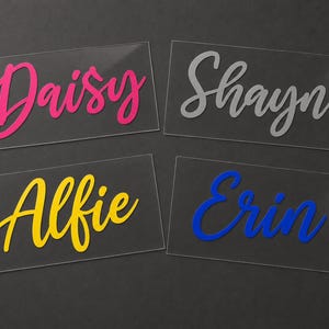 May include: Four clear acrylic nameplates with names in various colors. The names are Daisy in pink, Shayne in gray, Alfie in yellow, and Erin in blue. The names are in a cursive font.