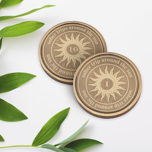 Op de afbeelding: Twee ronde houten onderzetters met een zonontwerp en de tekst "sober trips around the sun". E&eacute;n onderzetter heeft het nummer 10, de andere het nummer 1. Groene bladeren staan op de achtergrond.
