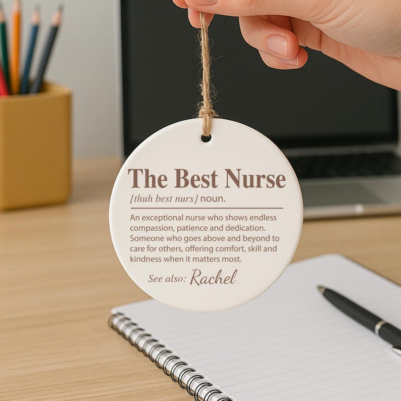 May include: A round, white ceramic ornament with the text "The Best Nurse" in brown lettering. The ornament includes a definition of a nurse and the name "Rachel". A brown string is attached for hanging.