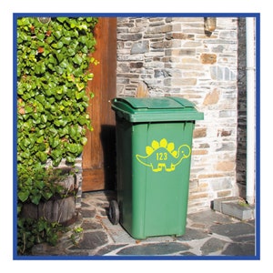 May include: Green recycling bin with a yellow dinosaur sticker and the number 123 on it. The bin is sitting on a stone patio in front of a brick wall.