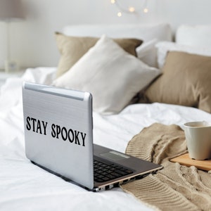 May include: A silver laptop computer with the text "STAY SPOOKY" on the lid. The laptop is on a white bed with a brown blanket and a white coffee mug.