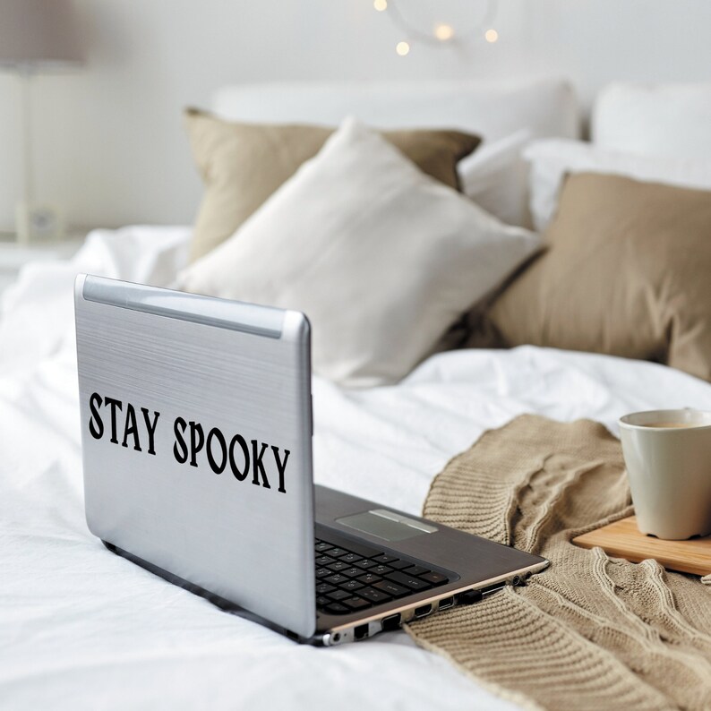 May include: A silver laptop computer with the text "STAY SPOOKY" on the lid. The laptop is on a white bed with a brown blanket and a white coffee mug.