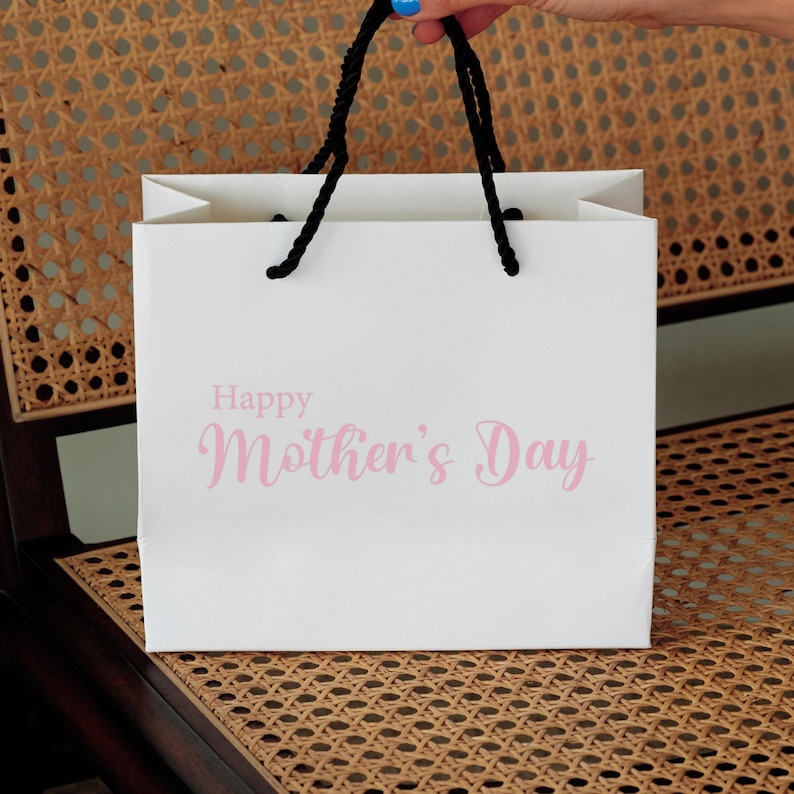 Puede incluir: Una bolsa de regalo blanca con asas negras y el texto "Happy Mother's Day" en letras rosas.