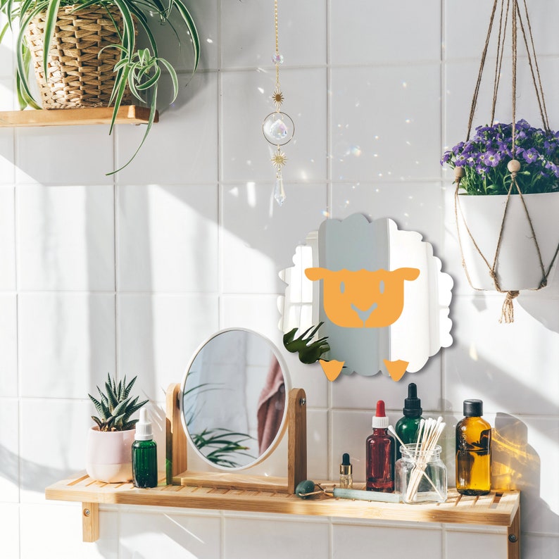 May include: A bathroom scene with a sheep-shaped mirror with an orange face and light blue body. A round mirror, plants, and various bottles are on a wooden shelf. A crystal suncatcher hangs from the ceiling.
