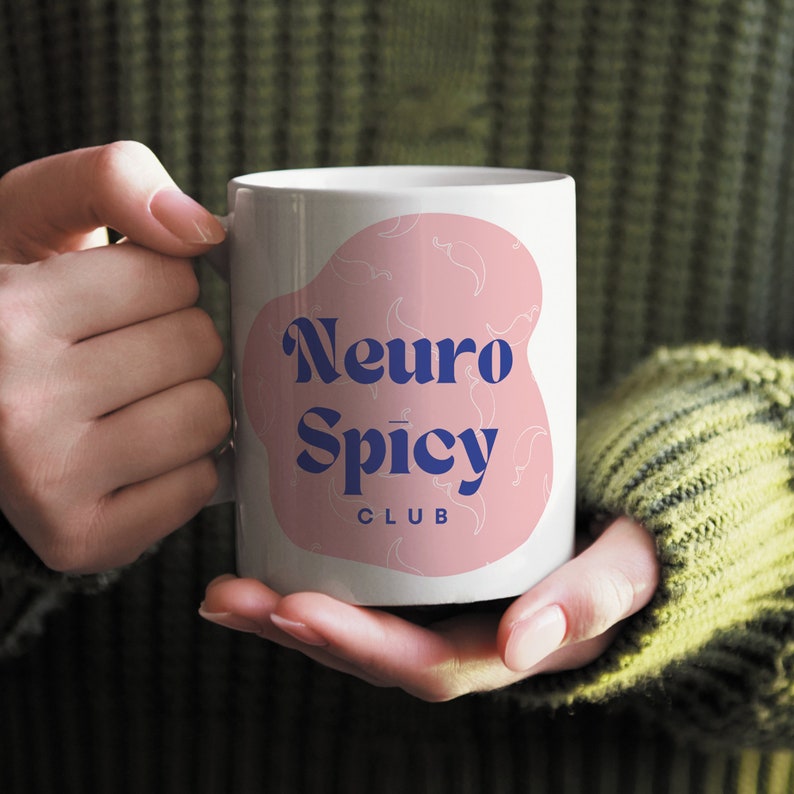 May include: White ceramic mug with a pink and white design. The mug has the text "Neuro Spicy Club" in blue lettering.