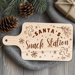 May include: A light-colored wooden cutting board with a handle, engraved with the words "SANTA'S Snack Station" in a decorative font. The board features gingerbread men, snowflakes, and other festive designs. Perfect for holiday entertaining.