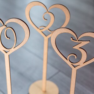 May include: Four wooden heart-shaped table numbers with a decorative swirl design. The numbers are 1, 2, 3, and 4.