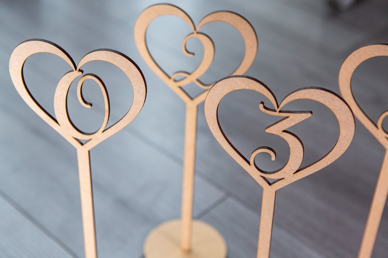 May include: Four wooden heart-shaped table numbers with a decorative swirl design. The numbers are 1, 2, 3, and 4.