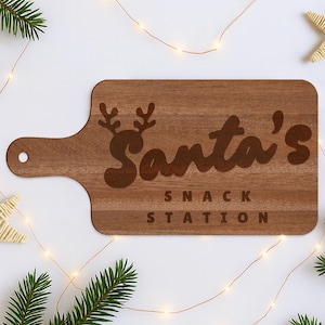 Op de afbeelding: Een bruine houten snijplank met handvat, gegraveerd met "Santa's Snack Station" en rendiergewei. Het bord is omgeven door kerstversieringen, waaronder dennentakken en lichtslingers.