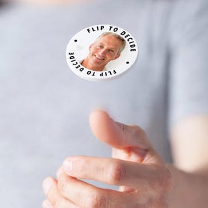May include: A white, round decision coin with the text "Flip to Decide" encircling a smiling man's face. The coin has black dots on the sides. The coin is being flipped in the air.