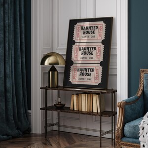 May include: A framed print featuring three vintage-style "Haunted House" tickets. The tickets are red and white with black text and a unique serial number. The print is displayed on a dark brown shelf with a gold lamp.