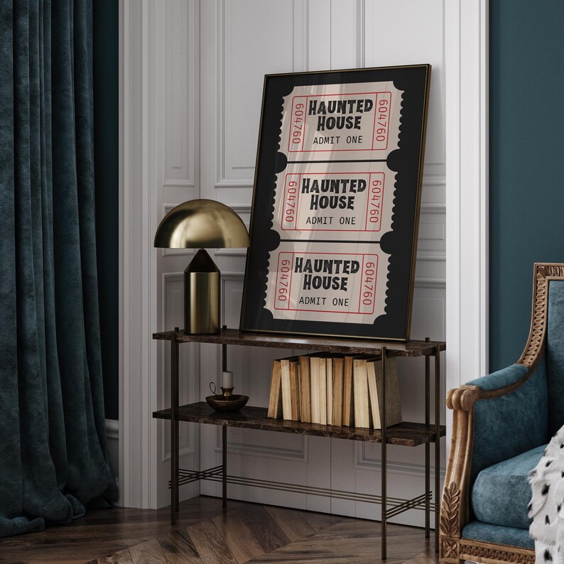 May include: A framed print featuring three vintage-style "Haunted House" tickets. The tickets are red and white with black text and a unique serial number. The print is displayed on a dark brown shelf with a gold lamp.