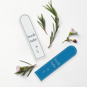 May include: Two white and blue bookmarks with the text "book babe" and a small flower design. The bookmarks are on a white background with sprigs of greenery and small pink flowers.