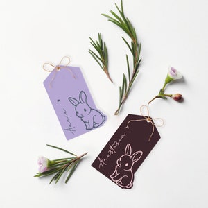May include: Two gift tags with bunny illustrations. The tag on the left is lavender with a gray bunny and the text "Annie" in gray. The tag on the right is brown with a pink bunny and the text "Anastasia" in white.