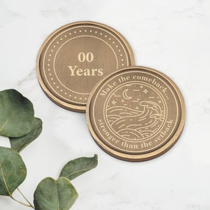 May include: Two round wooden coasters with gold lettering. The top coaster says "00 Years". The bottom coaster has a design of waves, a moon, and stars with the text "Make the comeback... stronger than the setback."