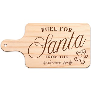 May include: A wooden cutting board with a handle, featuring the text "FUEL FOR Santa" in a decorative font. Below, it reads "FROM THE" and "AnySurname family". A gingerbread man graphic is on the right.