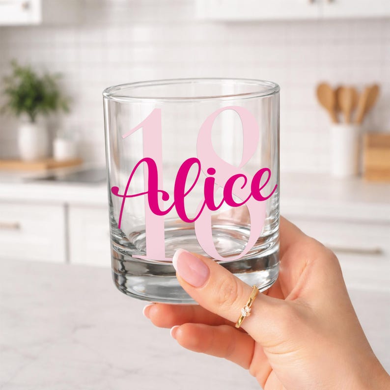 May include: A clear glass tumbler with the name "Alice" in bright pink script and the number "18" in light pink. The glass is held by a hand with a gold ring. The background is a blurred white kitchen.