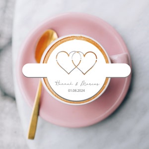 May include: A white ceramic coffee cup with a pink saucer. The coffee cup has a design of two intertwined hearts with the text "Hannah & Marcus" and the date "01.08.2024" printed on it.