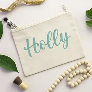 May include: A white canvas makeup bag with a zipper closure. The bag has the name "Holly" printed on it in teal glitter.