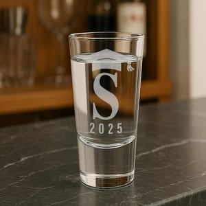 K&ouml;nnte beinhalten: Ein klares Schnapsglas, gef&uuml;llt mit Fl&uuml;ssigkeit, mit einem gro&szlig;en Buchstaben "S" und dem Jahr "2025" graviert. Das Glas hat ein Abschlusskappen-Design &uuml;ber dem Buchstaben. Das Glas steht auf einer dunklen Arbeitsplatte.