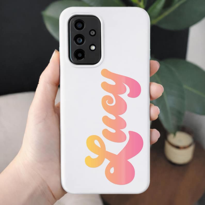 May include: White phone case featuring the name "Lucy" in a gradient orange to pink cursive font. The case has a camera module with three lenses.