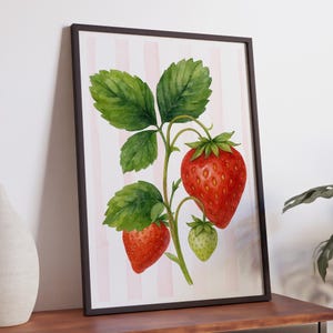 May include: Watercolor print of a strawberry plant with ripe red strawberries and green leaves. The artwork is framed in a dark brown frame and set against a background of pink and white vertical stripes. The print is displayed on a wooden surface.