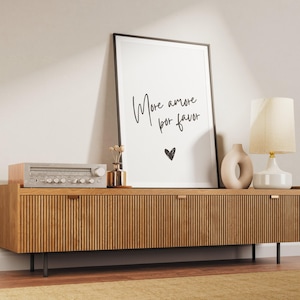 May include: A white framed print with the text "More amore por favor" and a small heart in black ink. The print is hanging on a wall above a wooden media console with two drawers.