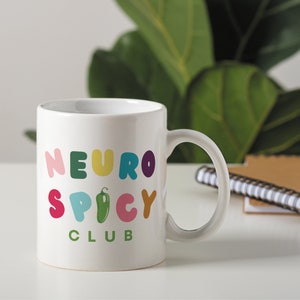 May include: White ceramic mug with a colorful graphic that says "Neuro Spicy Club" and a green pepper illustration.