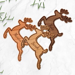 May include: Wooden reindeer ornaments in shades of brown, each with a name engraved: Marcus, Sarah, and Kate. The reindeer are leaping, with detailed antlers. The background is a white marble surface with green sprigs.