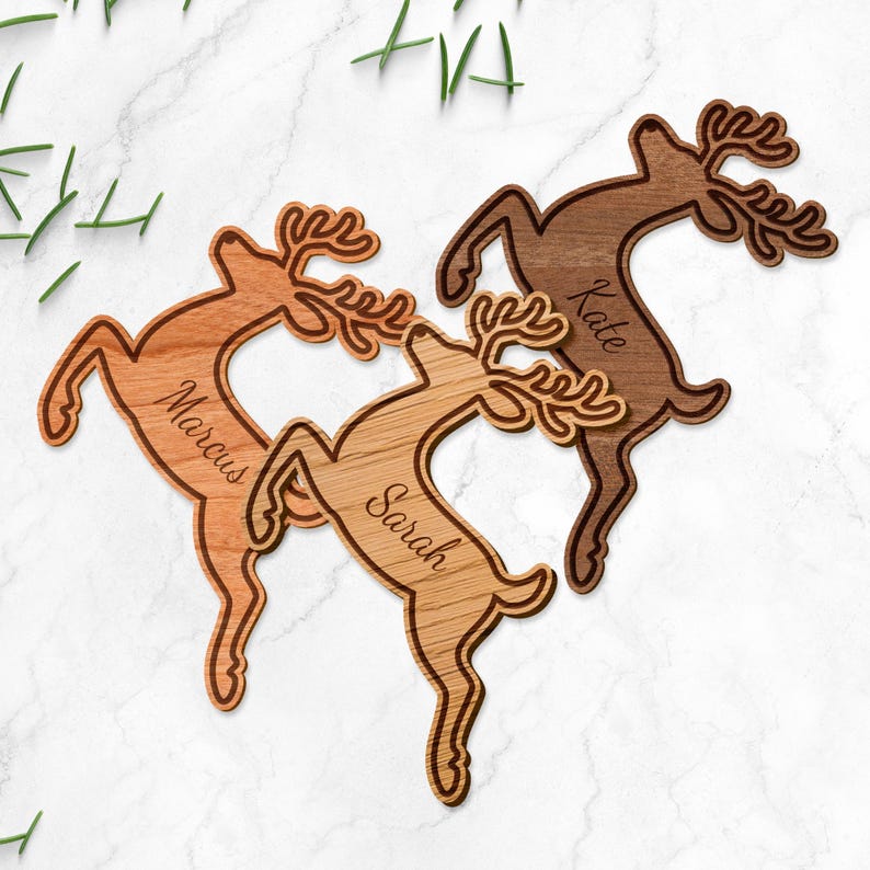 May include: Wooden reindeer ornaments in shades of brown, each with a name engraved: Marcus, Sarah, and Kate. The reindeer are leaping, with detailed antlers. The background is a white marble surface with green sprigs.