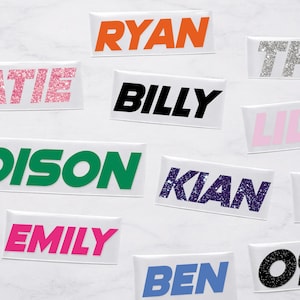 May include: A collection of name stickers in various colors and fonts. Names include Ryan, Billy, Emily, Kian, and others. Some names have glitter effects, while others are solid colors.