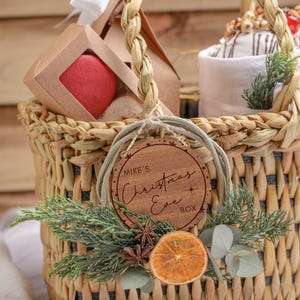 May include: A wicker basket filled with Christmas gifts, including a red macaron, a white towel, and a wooden ornament that says "Mike's Christmas Eve Box".