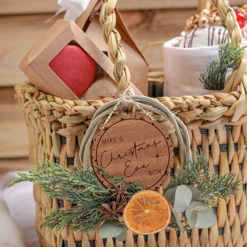 May include: A wicker basket filled with Christmas gifts, including a red macaron, a white towel, and a wooden ornament that says "Mike's Christmas Eve Box".