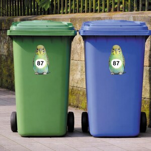 May include: Two large, wheeled trash bins, one green and one blue, stand side-by-side. Each bin has a sticker of a parakeet with the number 87. The bins are outdoors, with a fence and stone wall in the background.