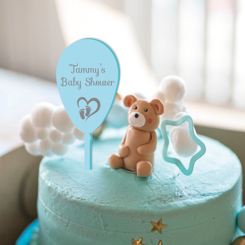May include: A blue and white cake with a blue balloon topper that says "Tammy's Baby Shower" with a heart and footprints. The cake is decorated with white clouds, a brown teddy bear, and blue stars.