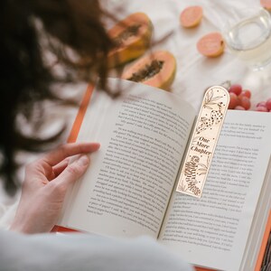Op de afbeelding: Een houten bladwijzer met de tekst "One More Chapter" en een ontwerp met een tijger en een luipaard. De bladwijzer is geplaatst in een open boek met oranje pagina's.