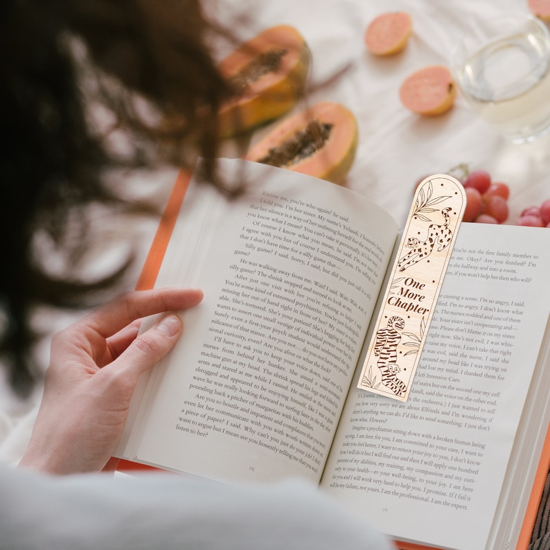 Op de afbeelding: Een houten bladwijzer met de tekst "One More Chapter" en een ontwerp met een tijger en een luipaard. De bladwijzer is geplaatst in een open boek met oranje pagina's.