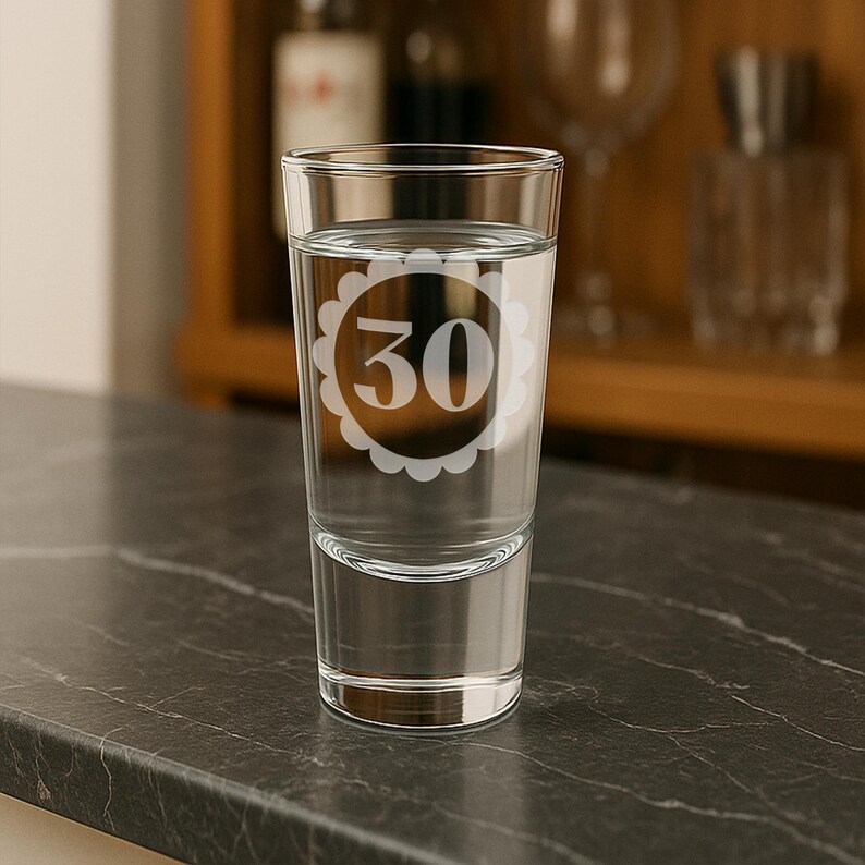 May include: A clear shot glass filled with water, featuring a white floral design around the number 30. The glass is on a dark countertop, with a blurred background of barware.