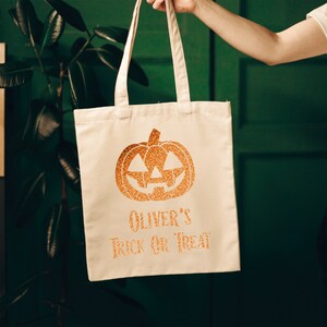 K&ouml;nnte beinhalten: Eine wei&szlig;e Stofftasche mit einem orangefarbenen Glitzerk&uuml;rbis und dem Text "Oliver's Trick or Treat".