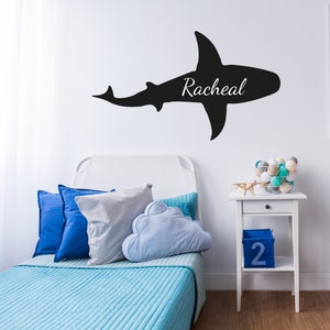 May include: A white bed with a blue comforter and pillows. A black shark silhouette wall decal with the name "Racheal" is on the wall above the bed. A white nightstand with a drawer and a blue box with the number "2" on it is next to the bed.