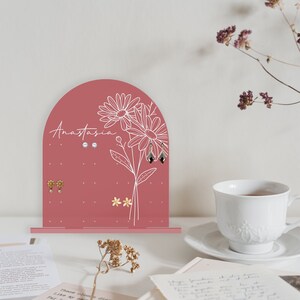 May include: A pink arched earring holder with a floral design and the name Anastasia written on it. The holder has multiple holes for earrings and is decorated with white flowers and leaves.