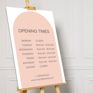 May include: A wooden easel displays a sign with "OPENING TIMES" in gray text. The sign has a peach-colored arch design and lists the shop's hours for each day of the week, including a phone number and website.