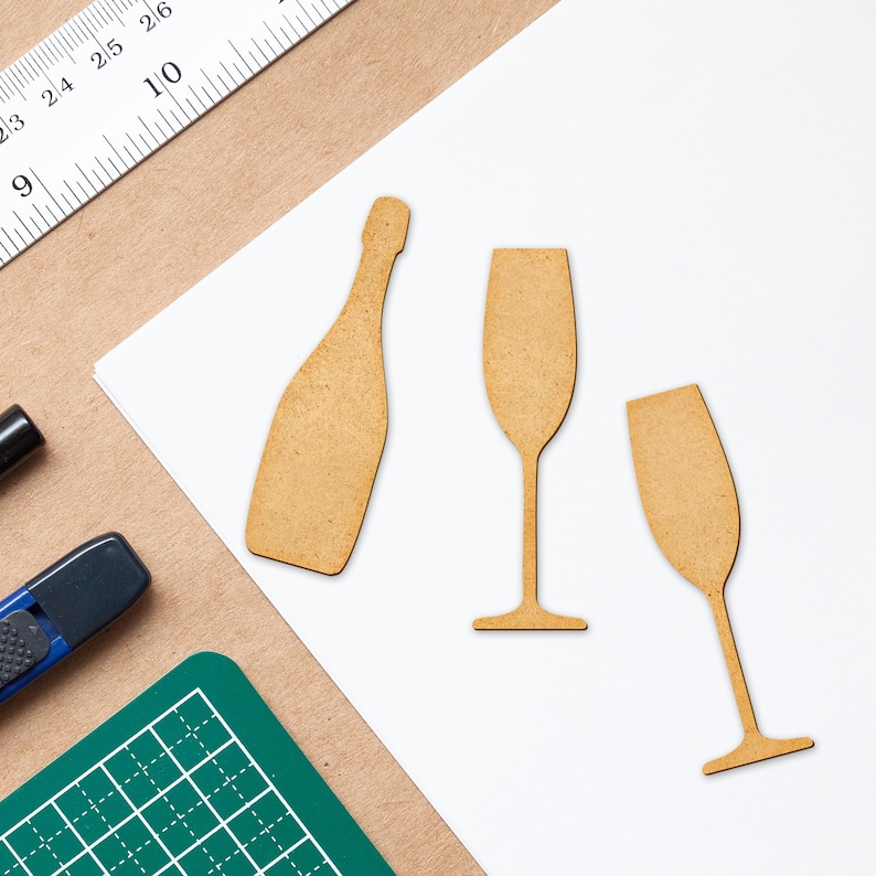 May include: Three wooden cutouts of a champagne bottle and two champagne flutes. The cutouts are light brown and are on a white surface.